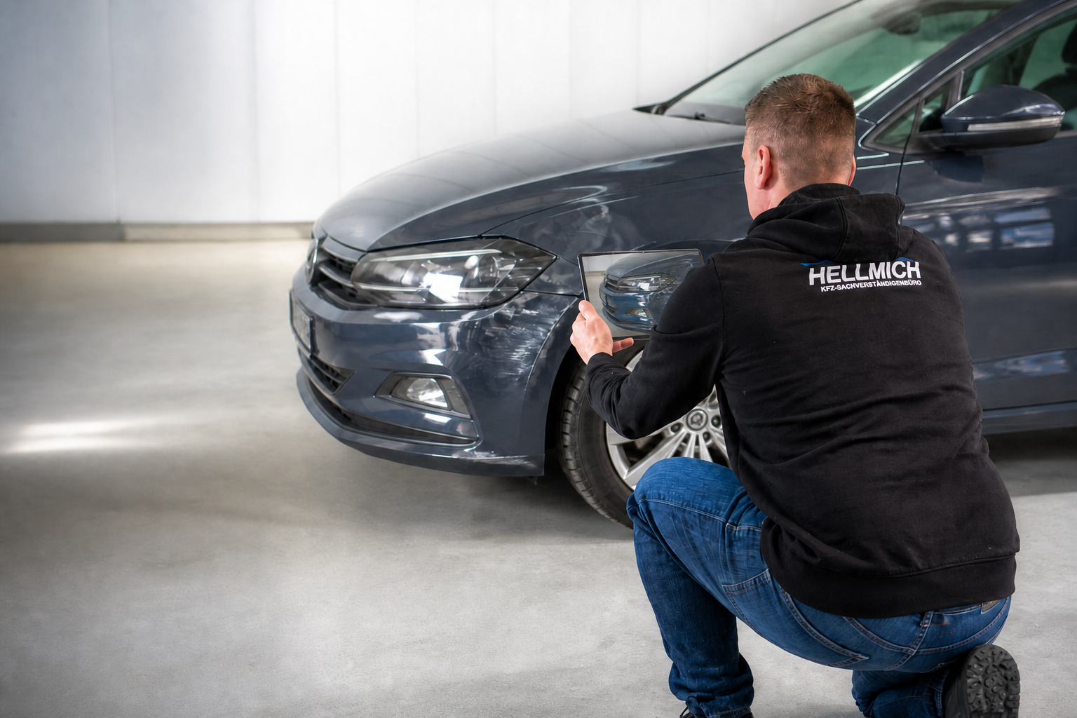 KFZ-Sachverständiger Siegfried Hellmich begutachtet kniend einen Unfallschaden am Fahrzeug mit Tablet